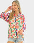 A woman wearing a colorful floral print button-up blouse with sheer puff sleeves and light-wash distressed denim shorts, posed against a solid light gray background.