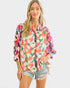 A woman modeling a long-sleeve button-up blouse with a vibrant multicolor floral print, paired with distressed denim shorts, against a solid light gray background.