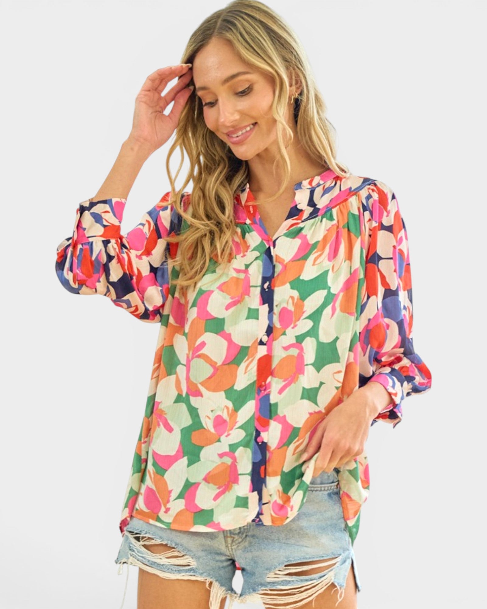 A woman wearing a long-sleeve button-up blouse with a vibrant multicolor floral print, paired with distressed denim shorts, against a solid light gray background.