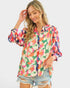 A woman wearing a long-sleeve button-up blouse with a vibrant multicolor floral print, paired with distressed denim shorts, against a solid light gray background.