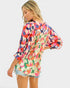A woman wearing a colorful abstract floral print blouse with three-quarter sleeves and a gathered back, paired with denim shorts, shown from a side-back angle against a light gray background.