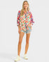 A woman modeling a colorful, long-sleeved button-up shirt with a vibrant abstract floral pattern, paired with denim shorts and white sneakers, set against a plain light gray background.