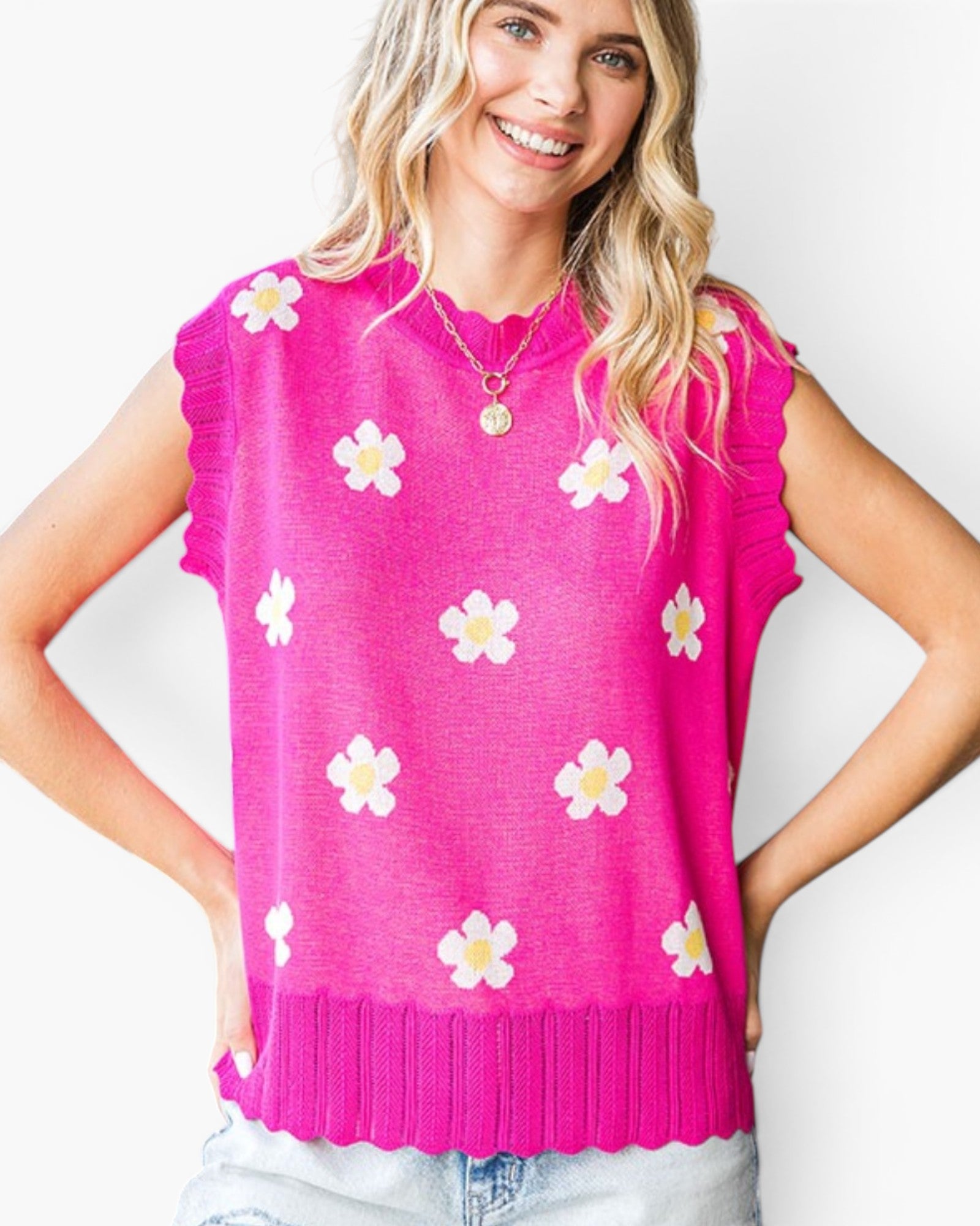 A woman wearing a bright pink sleeveless knit top with white and yellow flower motifs, paired with light blue jeans, against a plain white background.