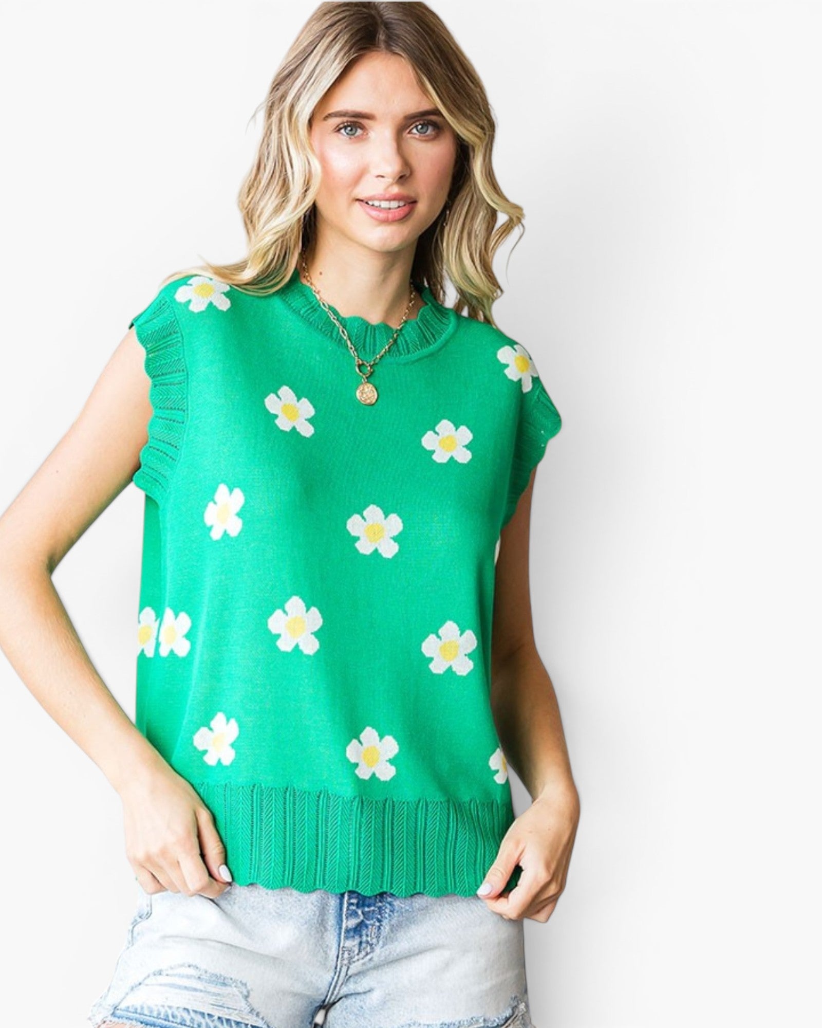 A woman wearing a bright green sleeveless knit top with a white and yellow daisy pattern, paired with light-wash denim shorts, against a plain white background.