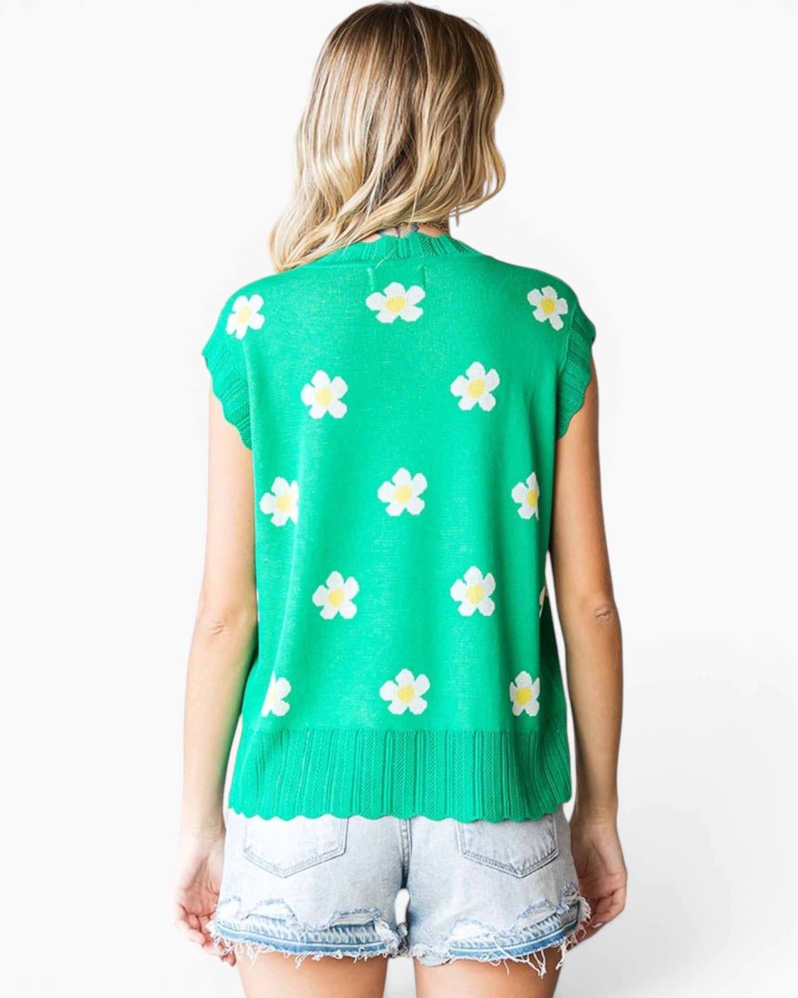A woman shown from the back wearing a bright green sleeveless knit top with a white and yellow daisy pattern, paired with light-wash denim shorts, against a plain white background.