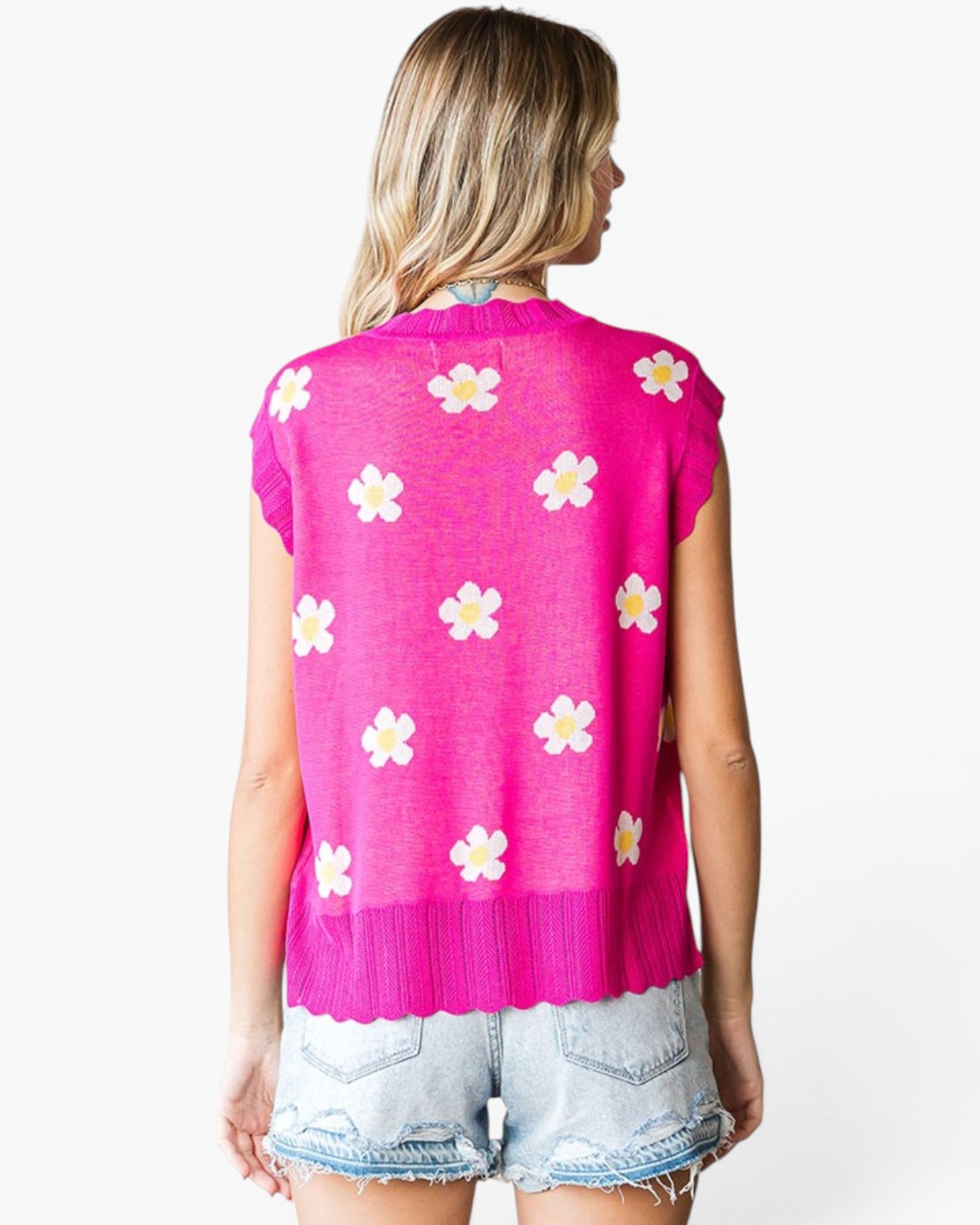 A woman shown from the back wearing a bright pink sleeveless knit top with a scalloped hem and an allover white daisy pattern, paired with light-wash denim shorts against a plain white background.