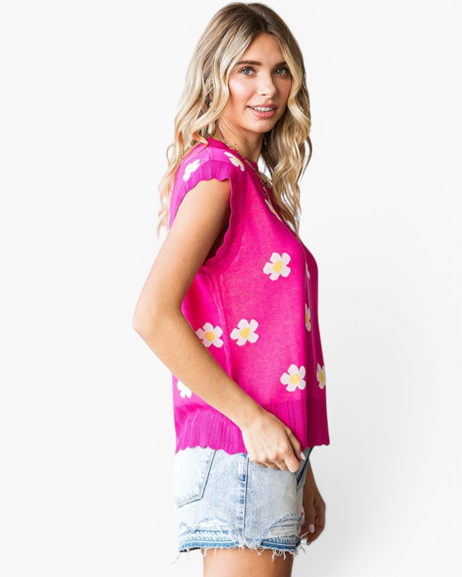 A woman modeling a bright pink sleeveless knit top with white and yellow daisy flower prints, paired with light-wash denim shorts, against a plain white background.