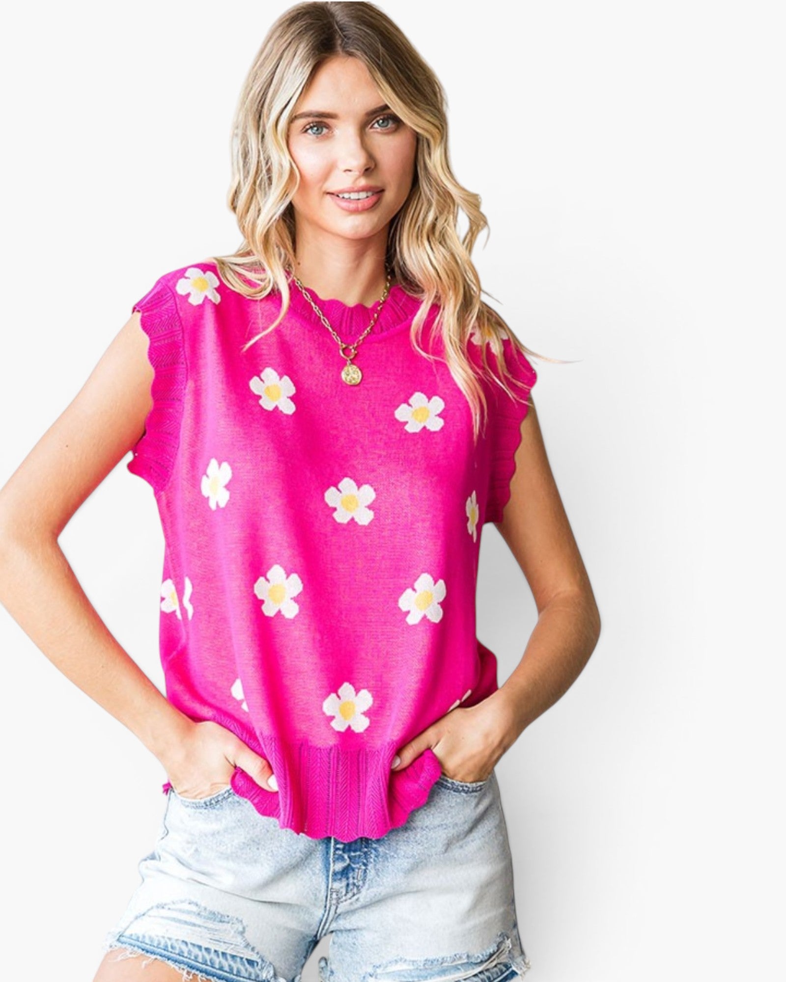 A woman wearing a bright pink sleeveless knit top with a white and yellow daisy pattern, paired with light-wash denim shorts, against a plain white background.