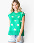 A woman wearing a bright green short-sleeve knit top with white daisy patterns and a fringed hem, paired with light-wash denim shorts, against a plain white background.