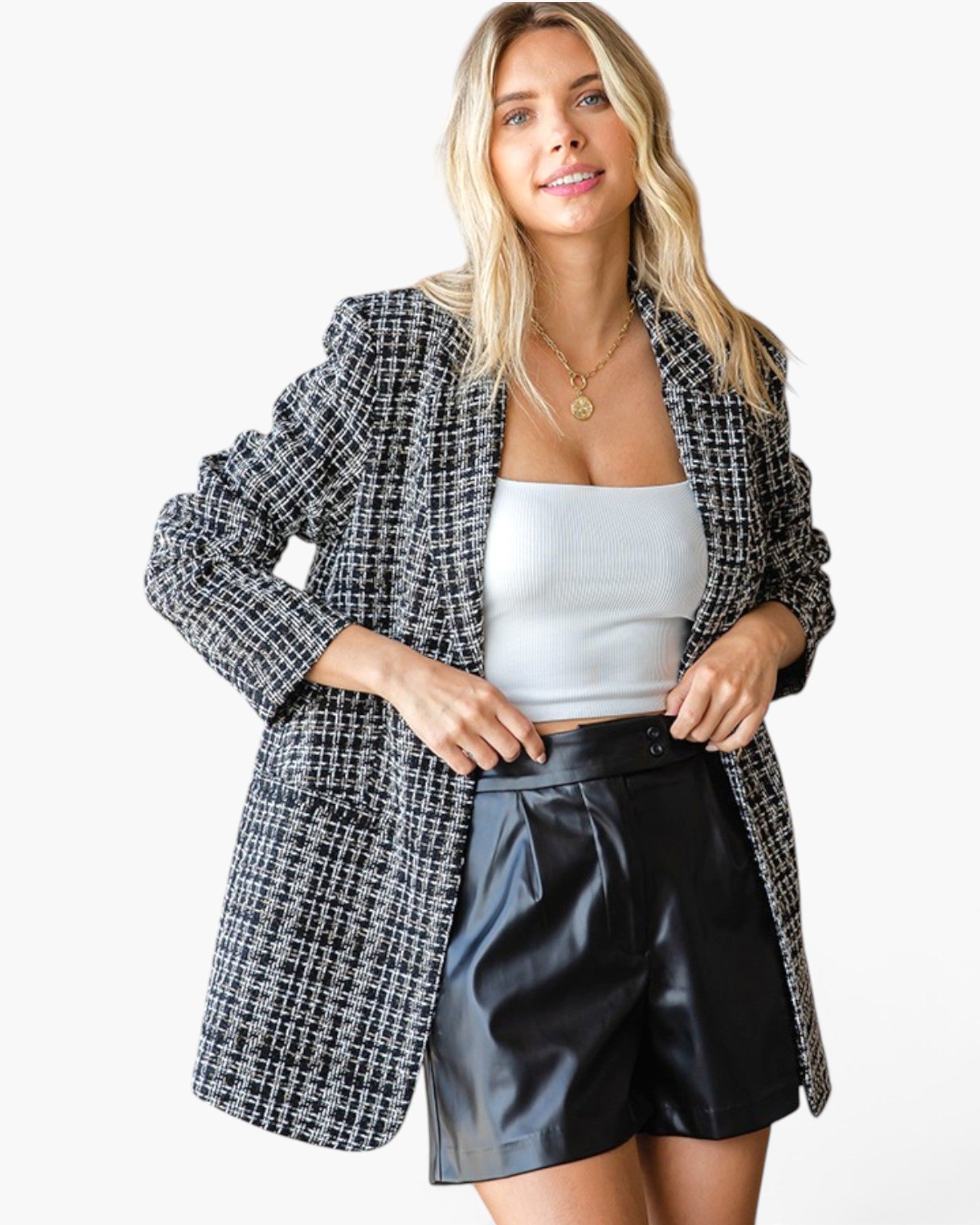 A woman modeling an oversized black and white houndstooth blazer over a white ribbed tank top and black faux-leather shorts, against a plain white studio background.