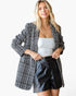 A woman modeling an oversized black and white houndstooth blazer over a white ribbed tank top and black faux-leather shorts, against a plain white studio background.