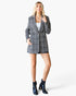 A woman modeling a black and white plaid blazer with black leather shorts and black ankle boots, standing against a light gray studio background.
