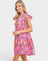 A woman modeling a tiered short dress with a vibrant abstract print in pink, orange, purple, and teal on a light gray background.