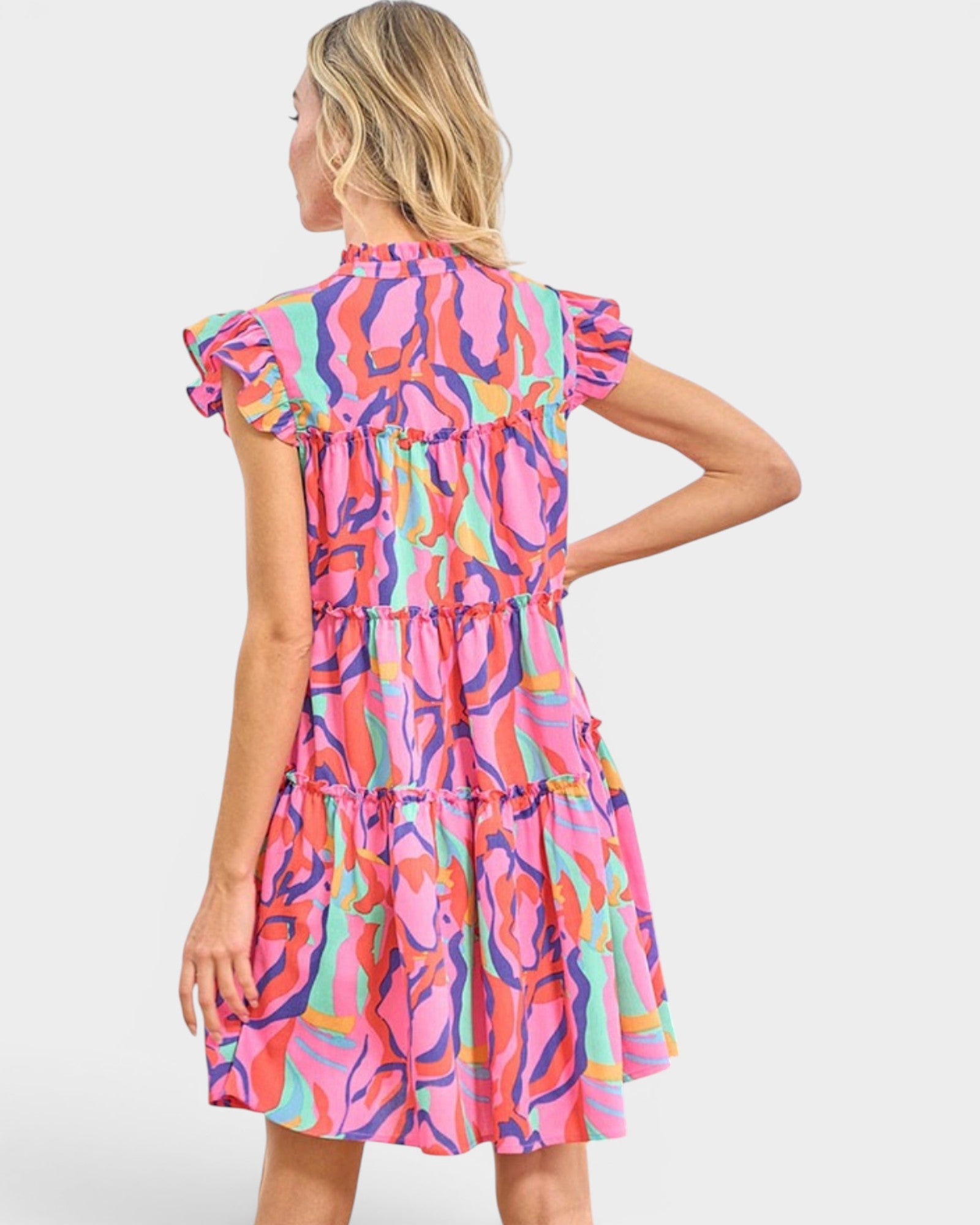 A woman shown from the back wearing a tiered short-sleeve dress with a bright abstract print in pink, orange, purple, and teal on a light gray studio background.