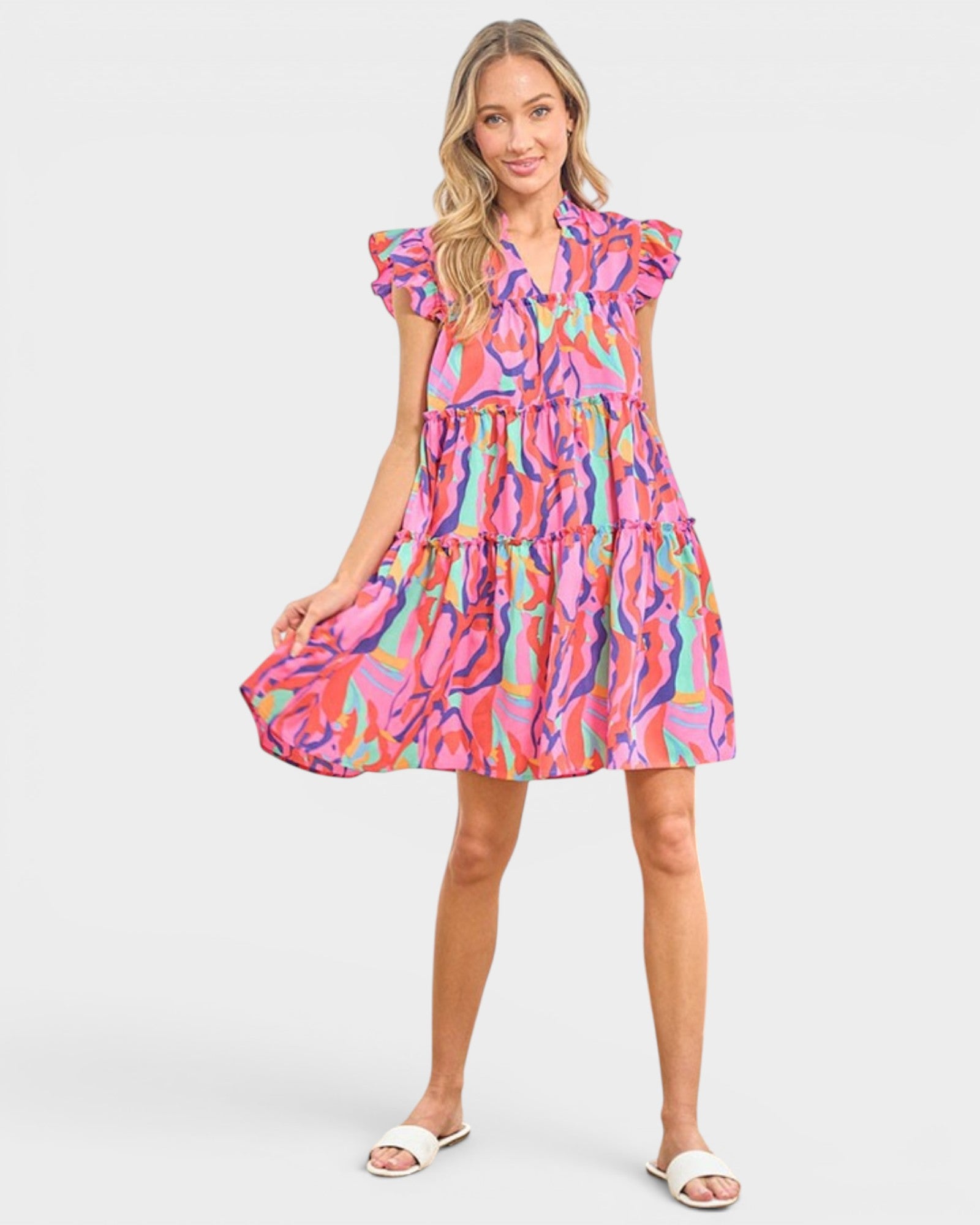 A woman modeling a short tiered dress with a vibrant abstract multicolor print in pink, orange, blue, and yellow, paired with white slide sandals against a light gray studio background.