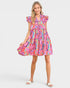 A woman modeling a short tiered dress with a vibrant abstract multicolor print in pink, orange, blue, and yellow, paired with white slide sandals against a light gray studio background.