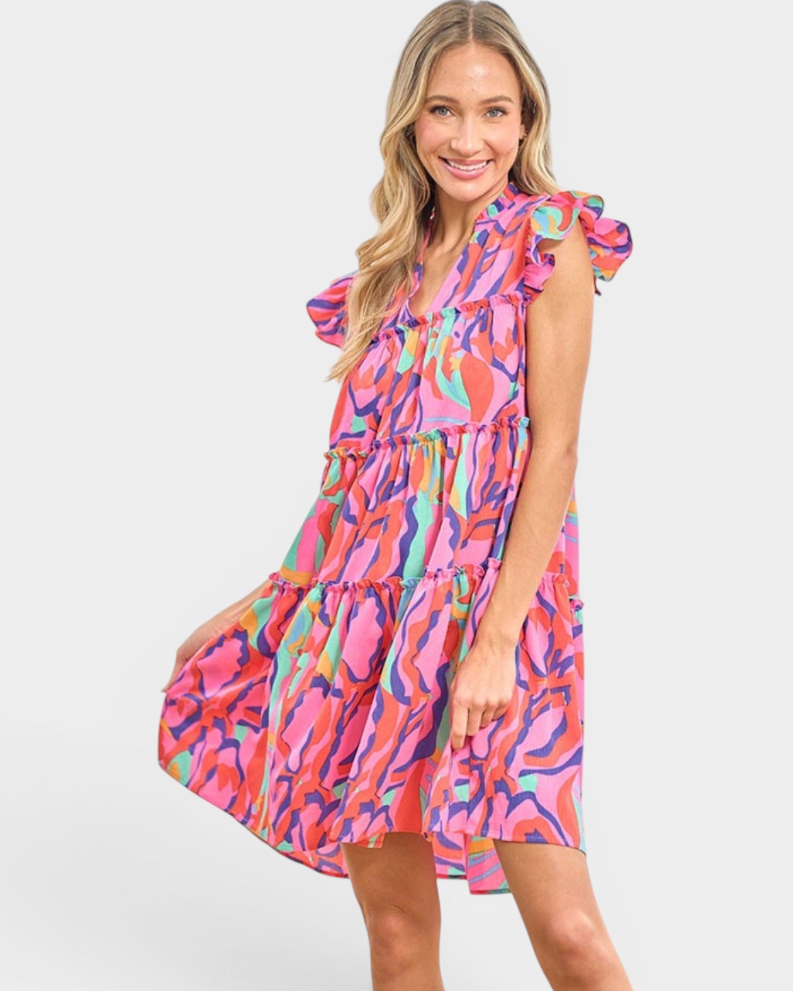 A woman modeling a tiered mini dress with a vibrant multicolor abstract print in pink, orange, purple, and blue, set against a plain light gray background.