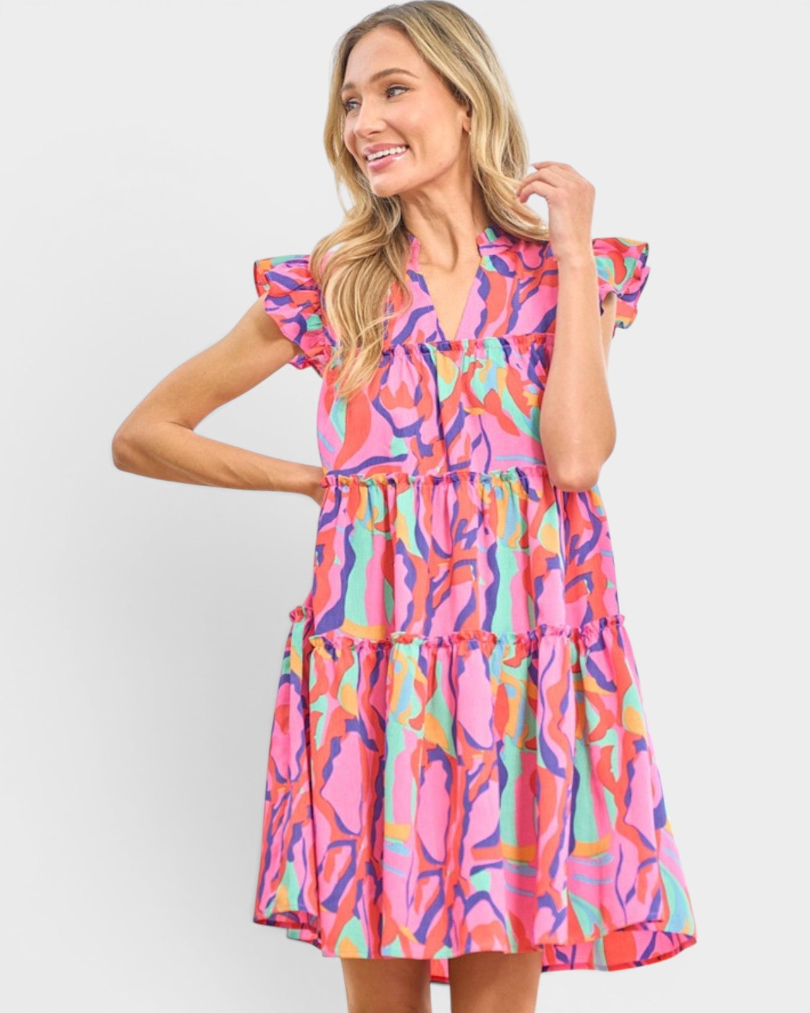 A woman modeling a tiered V-neck dress with a vibrant abstract print in pink, orange, yellow, green, and blue, set against a plain light gray background.