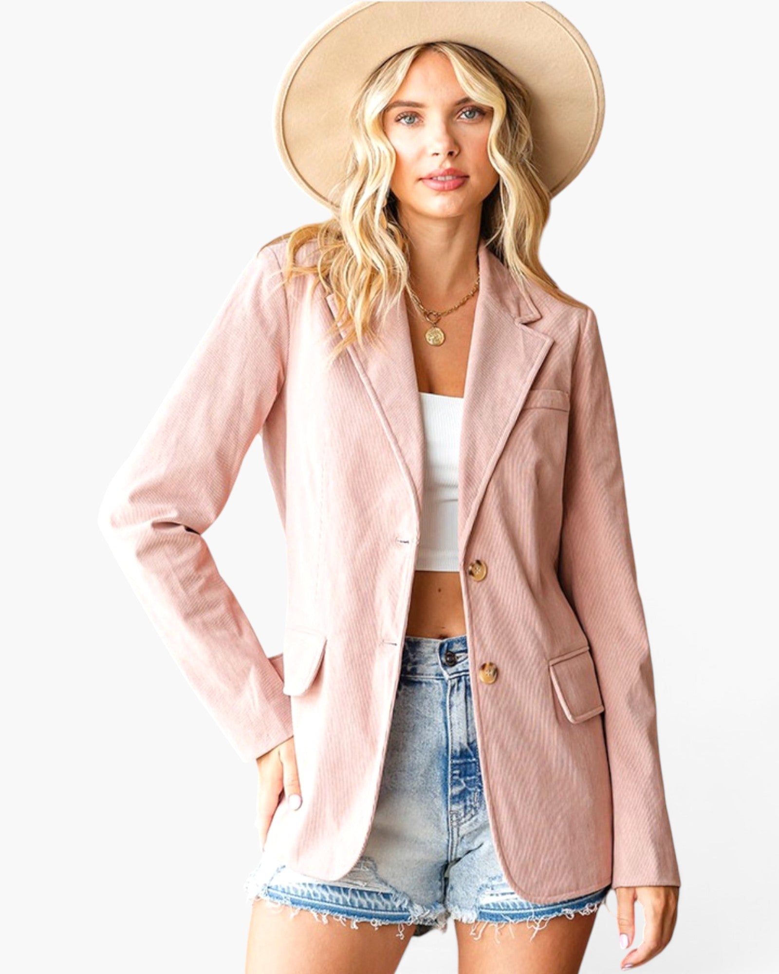 A woman modeling a light pink corduroy blazer, styled with a beige wide-brim hat, a white crop top, and denim shorts against a plain white background.