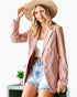 A woman models a dusty pink tailored blazer with gold buttons, styled with a white crop top, denim shorts, a beige wide-brim hat, and gold jewelry against a clean white background.