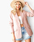 A woman modeling a dusty pink blazer with a white crop top and denim shorts, wearing a beige wide-brim hat against a plain white background.