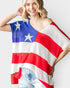 A woman wearing a loose, off-the-shoulder knit poncho top with an American flag pattern of red and white stripes and a blue field with white stars, paired with light-wash denim shorts, against a light gray background.