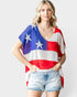 A woman modeling a short-sleeve V-neck knit top with an American flag pattern featuring red, white, and blue stripes and white stars, paired with light-wash denim shorts against a light gray background.