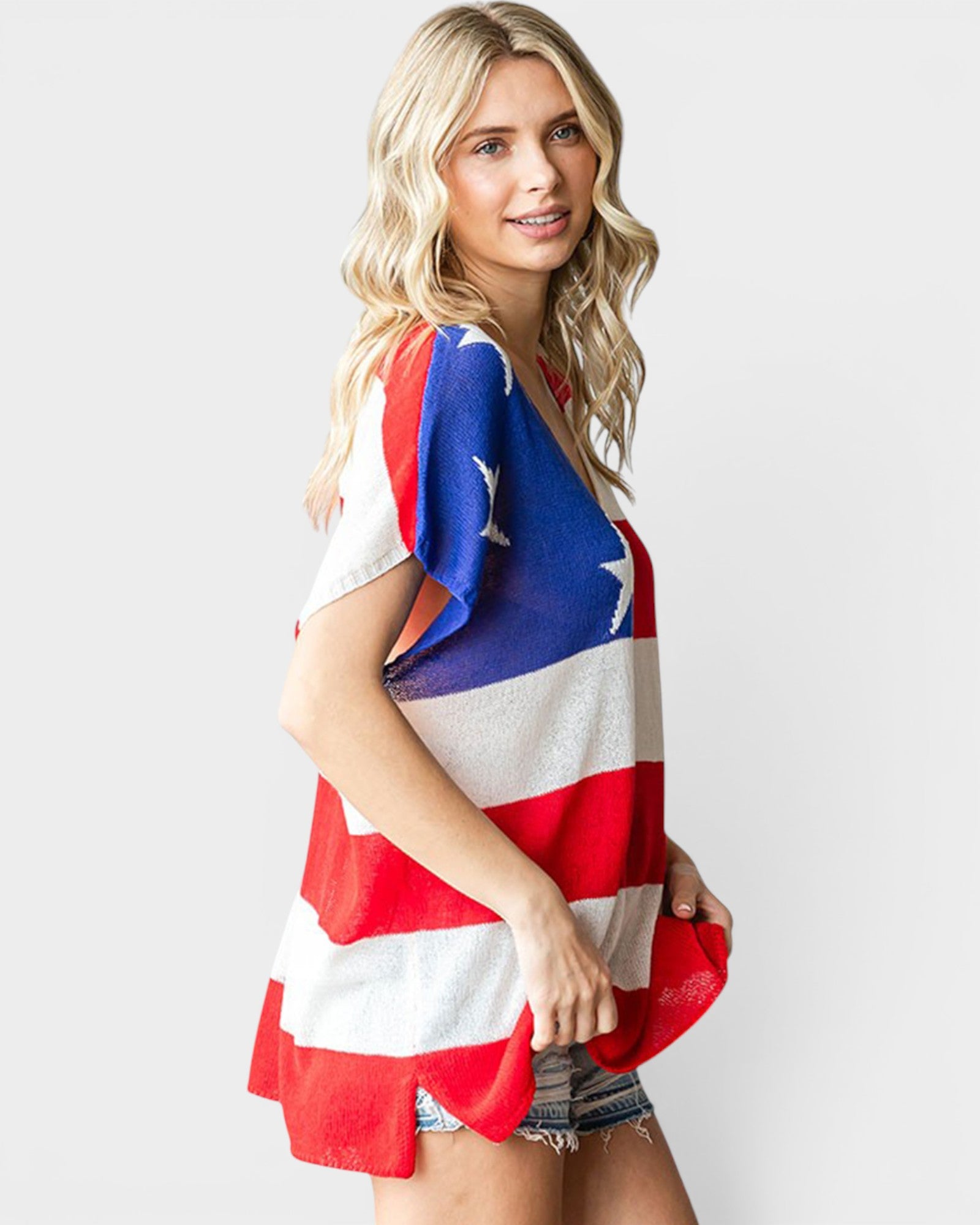 A woman modeling a short-sleeve V-neck top with an American flag pattern featuring red and white horizontal stripes and a blue section with white stars, against a light gray studio background.