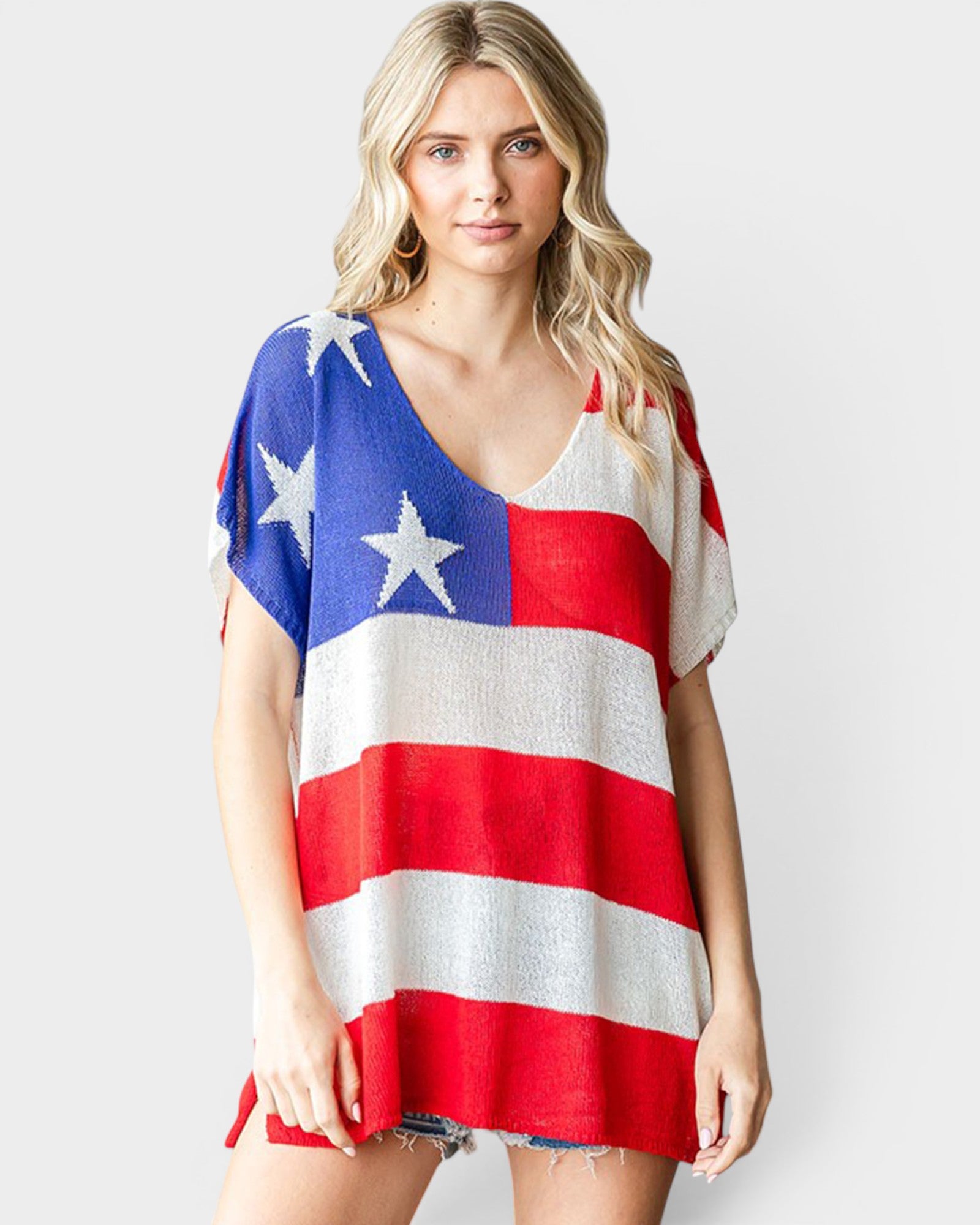 A woman modeling a short-sleeve V-neck tunic top with an American flag pattern featuring red and white stripes and white stars on a blue field, set against a plain light gray background.