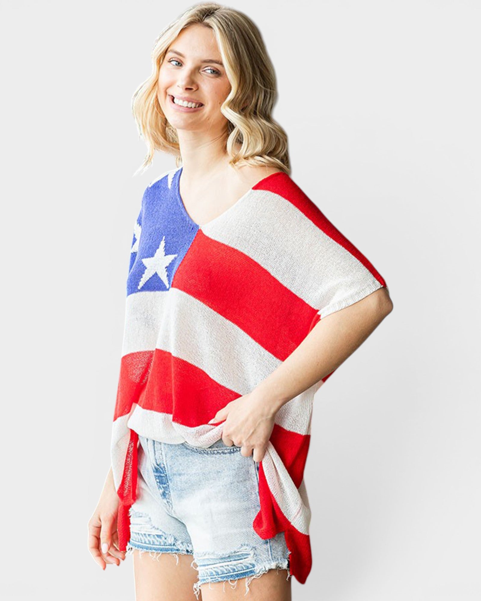 A woman modeling a red, white, and blue knit poncho top with an American flag pattern, paired with light-wash denim shorts, against a light gray studio background.