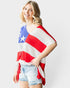 A woman modeling a red, white, and blue knit poncho top with an American flag pattern, paired with light-wash denim shorts, against a light gray studio background.