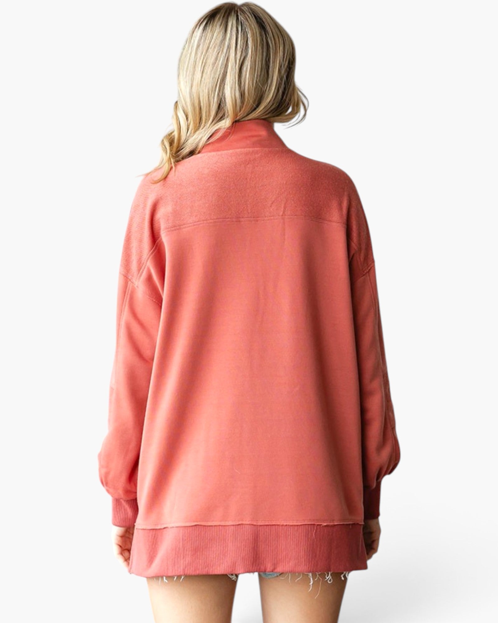A woman viewed from the back wearing an oversized long-sleeve sweatshirt in a muted coral pink color, set against a plain light gray studio background.