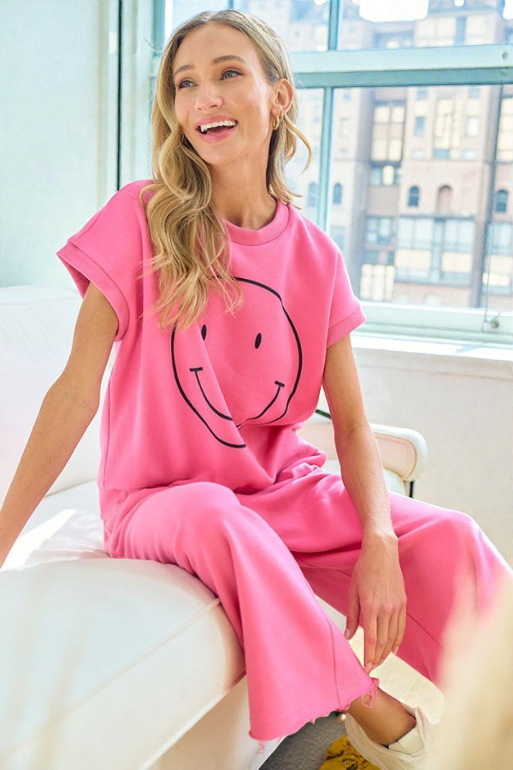 Woman wearing a pink outfit with a smiley face design, sitting in a bright room.