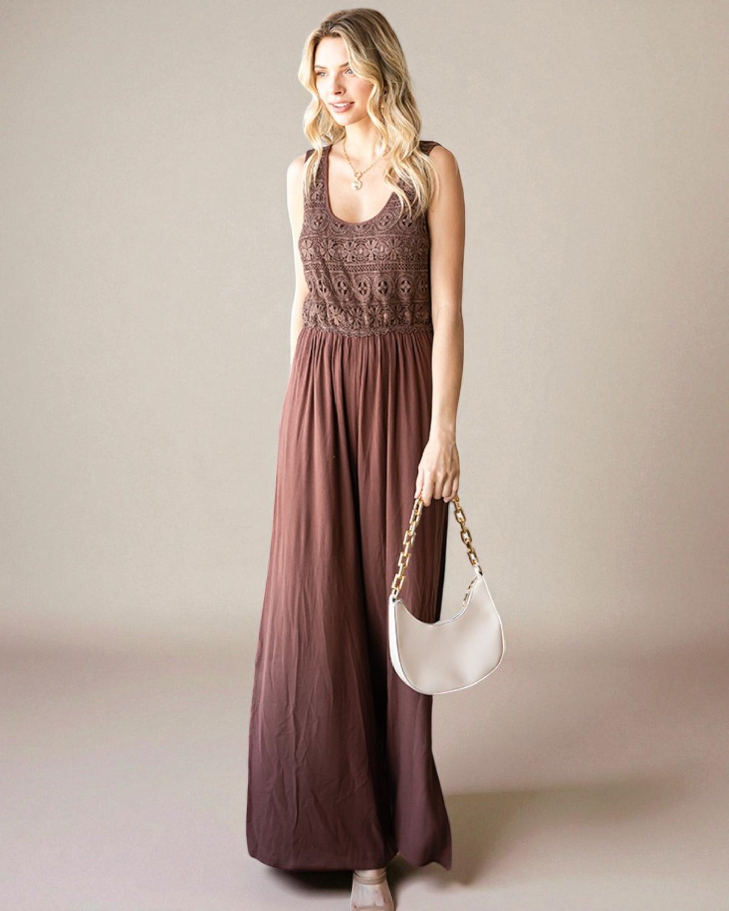 Woman wearing a long brown dress holding a white handbag against a plain background