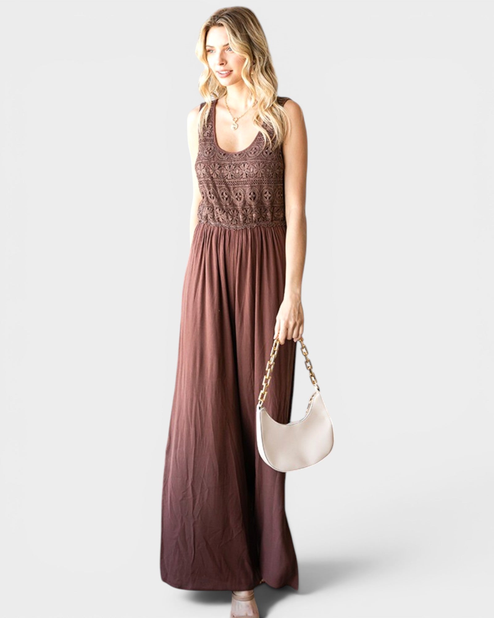 Woman wearing a long brown dress with a lace top, holding a white handbag on a light gray background.