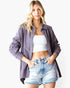 A woman modeling a loose, long-sleeve button-up shirt in a muted dusty purple or mauve color, worn open over a white crop top and light-wash denim shorts, against a plain white studio background.