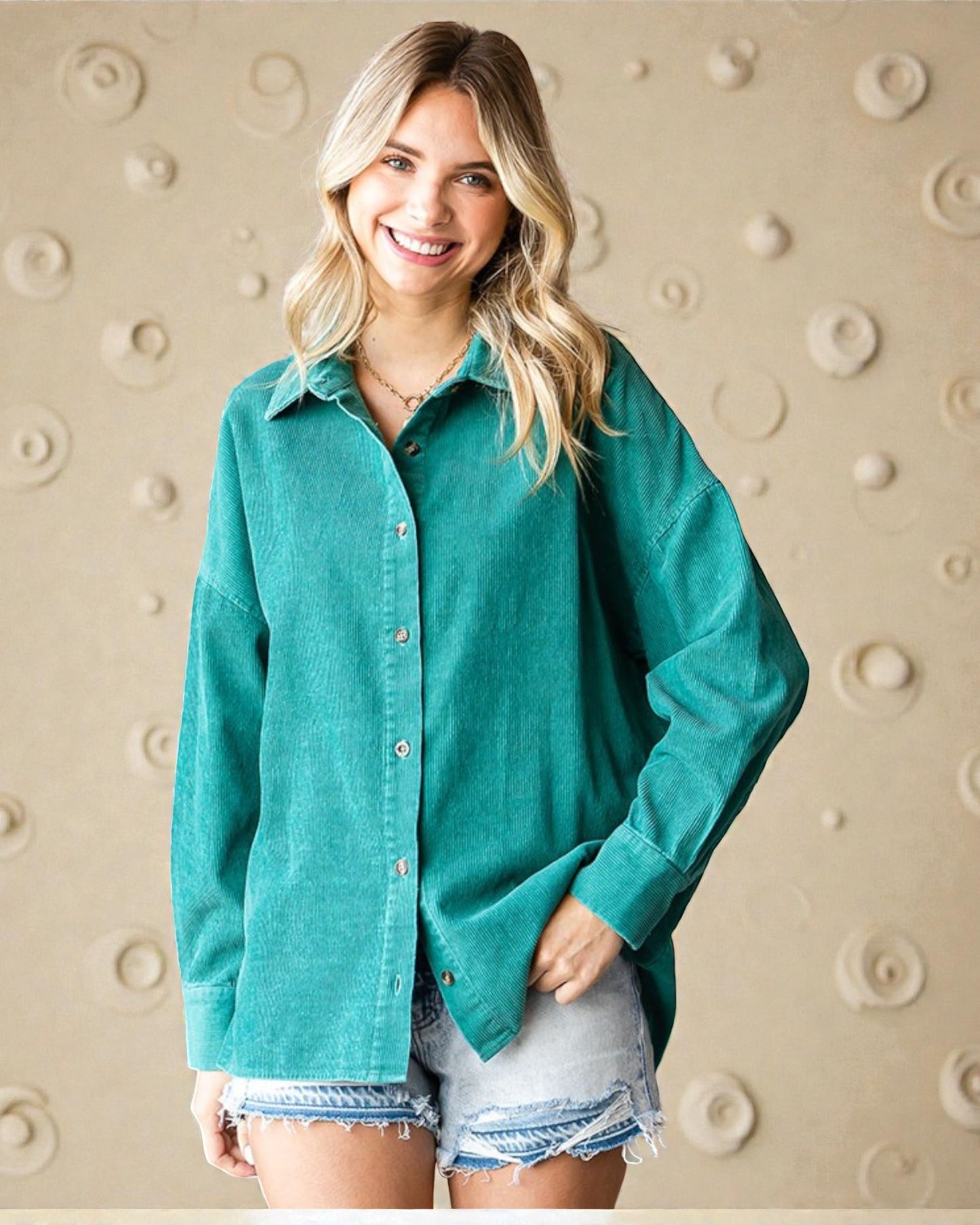 Woman wearing a teal button-up shirt and denim shorts against a beige wall with circular patterns.
