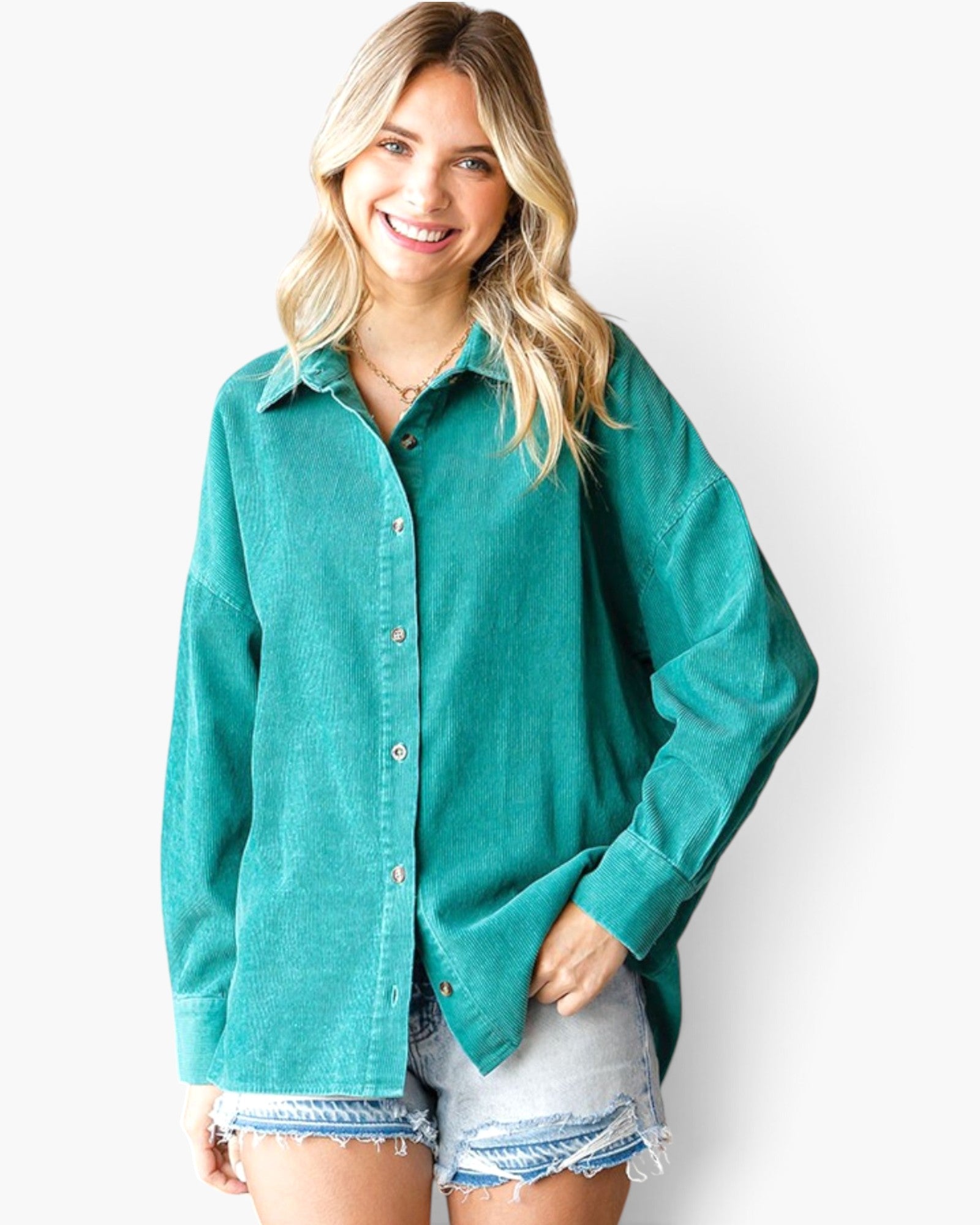 A woman wearing a teal green long-sleeve button-down shirt and light-wash denim shorts, standing against a plain white studio background.