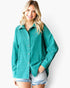 A woman wearing a teal green long-sleeve button-down shirt and light-wash denim shorts, standing against a plain white studio background.