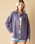 Woman wearing a purple denim jacket over a white top and denim shorts on a beige background
