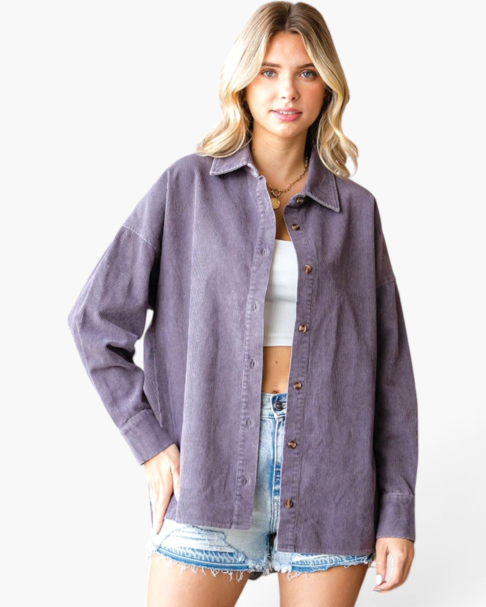 A woman modeling an oversized, long-sleeve button-up shirt in a washed, muted purple or mauve color, worn open over a white crop top and denim shorts, against a plain white studio background.