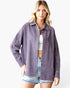 A woman modeling an oversized, long-sleeve button-up shirt in a washed, muted purple or mauve color, worn open over a white crop top and denim shorts, against a plain white studio background.
