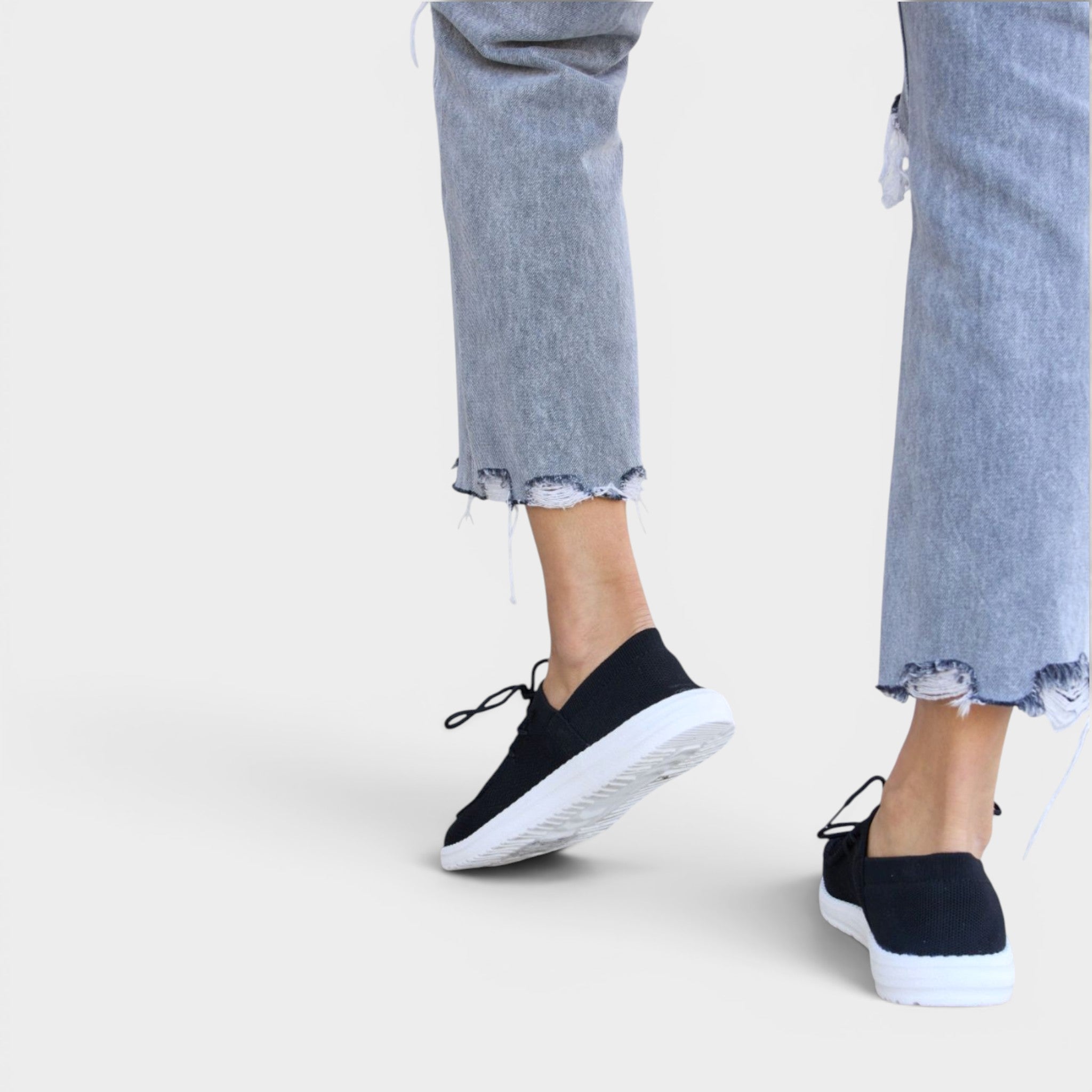 Black slip-on sneakers worn with light blue jeans on a white background