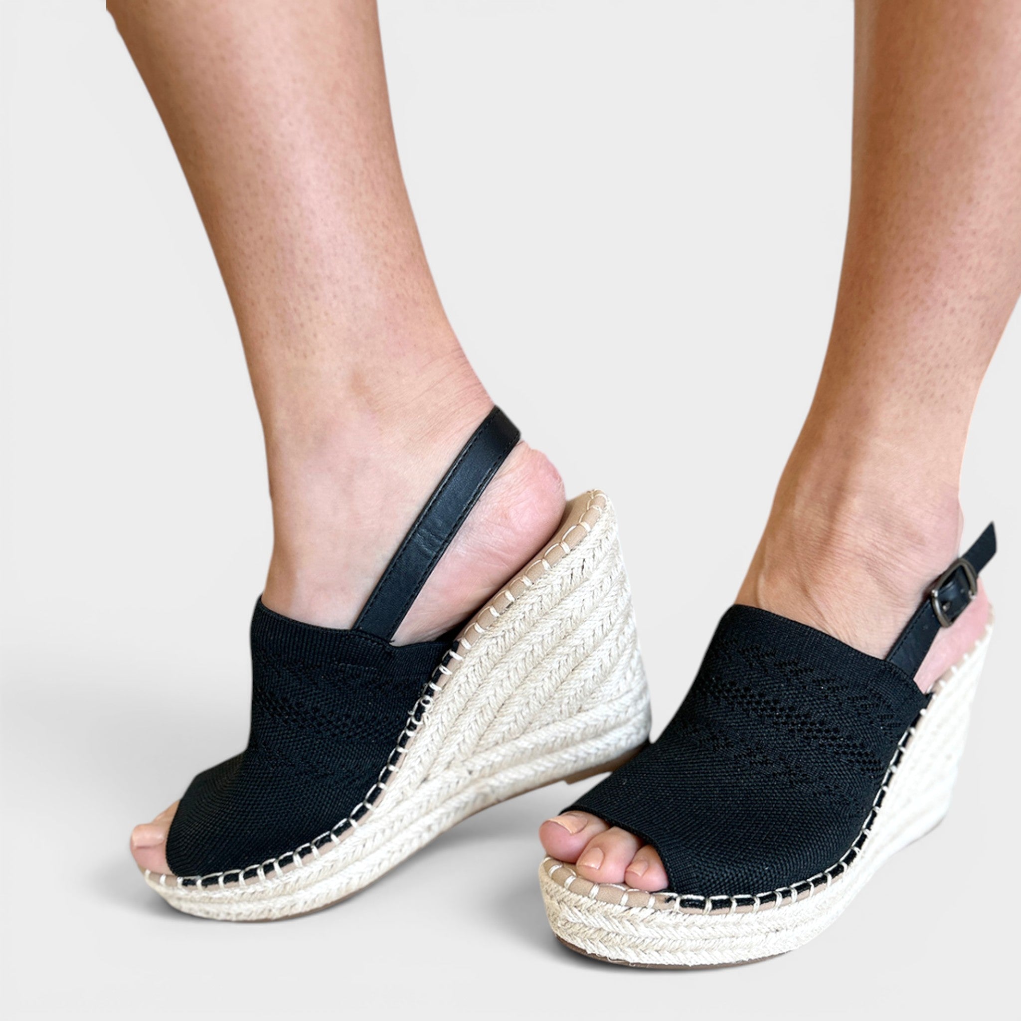 A pair of women's black woven wedge sandals with braided jute espadrille-style wedge heels and ankle straps, shown worn on a model's feet against a light gray studio background.