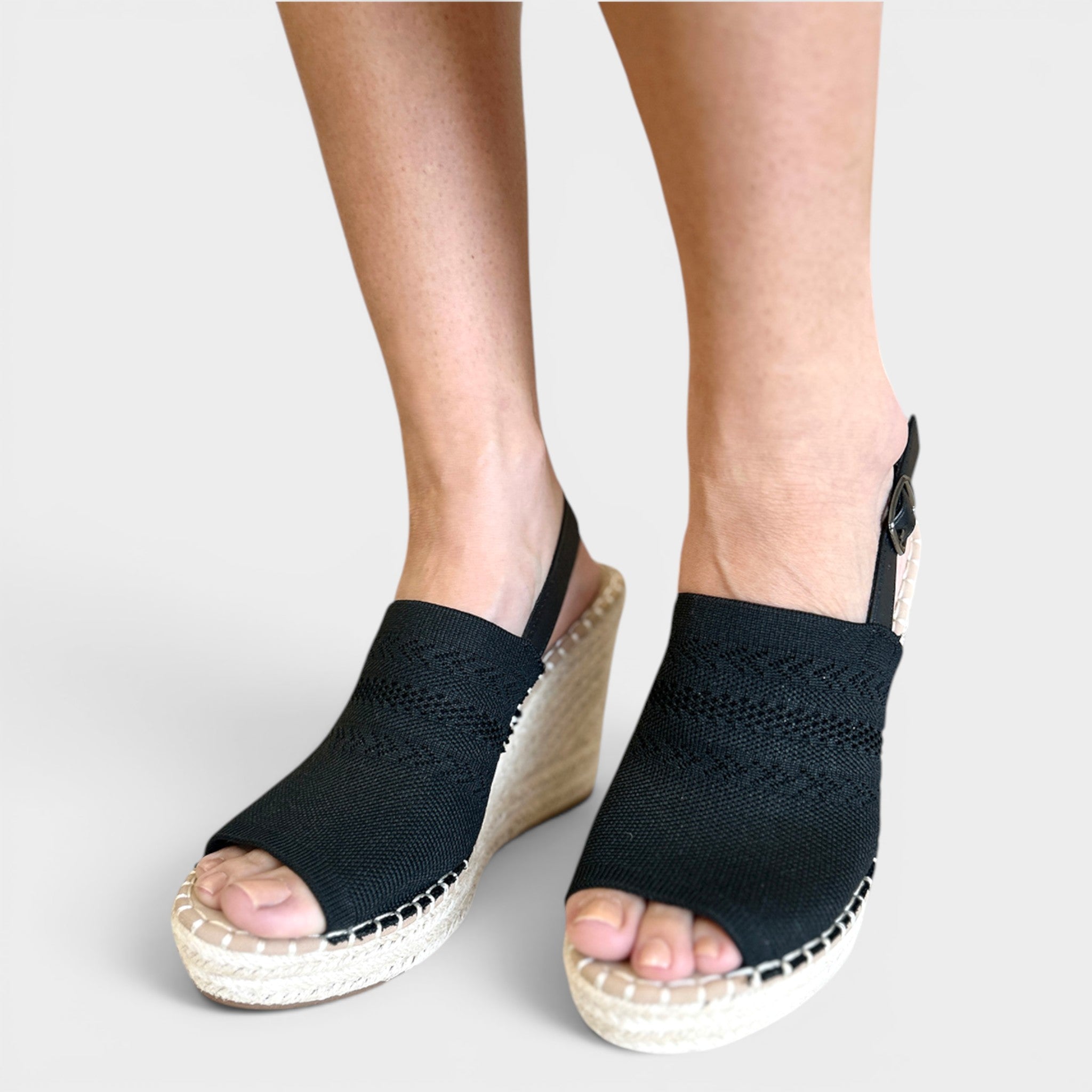 A pair of women's black woven espadrille wedge sandals with open toes, shown on a model's feet against a light gray background.