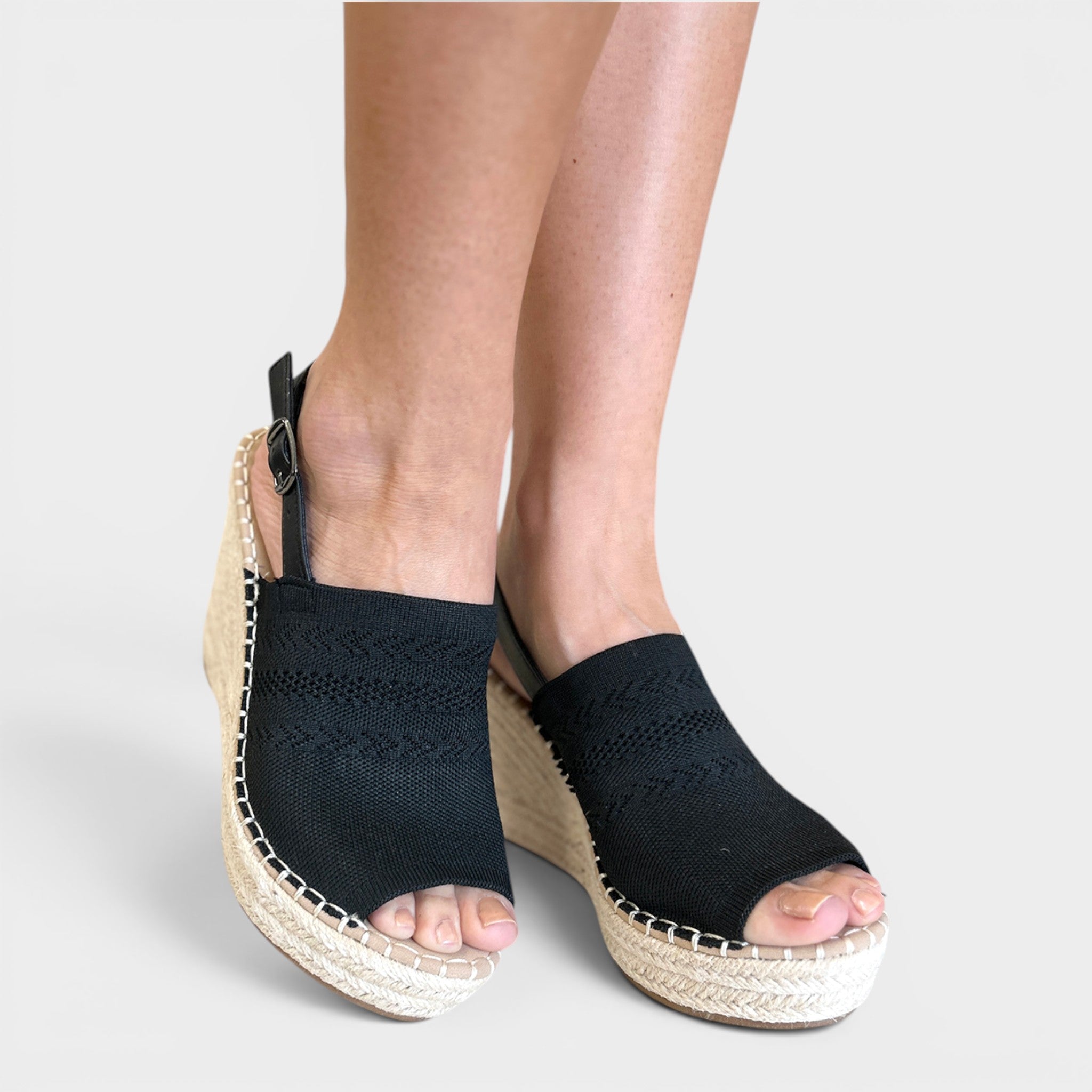 A pair of women's black woven espadrille wedge sandals with open toes and ankle straps, shown worn on a model's feet against a light gray background.
