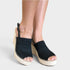 A pair of women's black woven espadrille wedge sandals with open toes and ankle straps, shown worn on a model's feet against a light gray background.