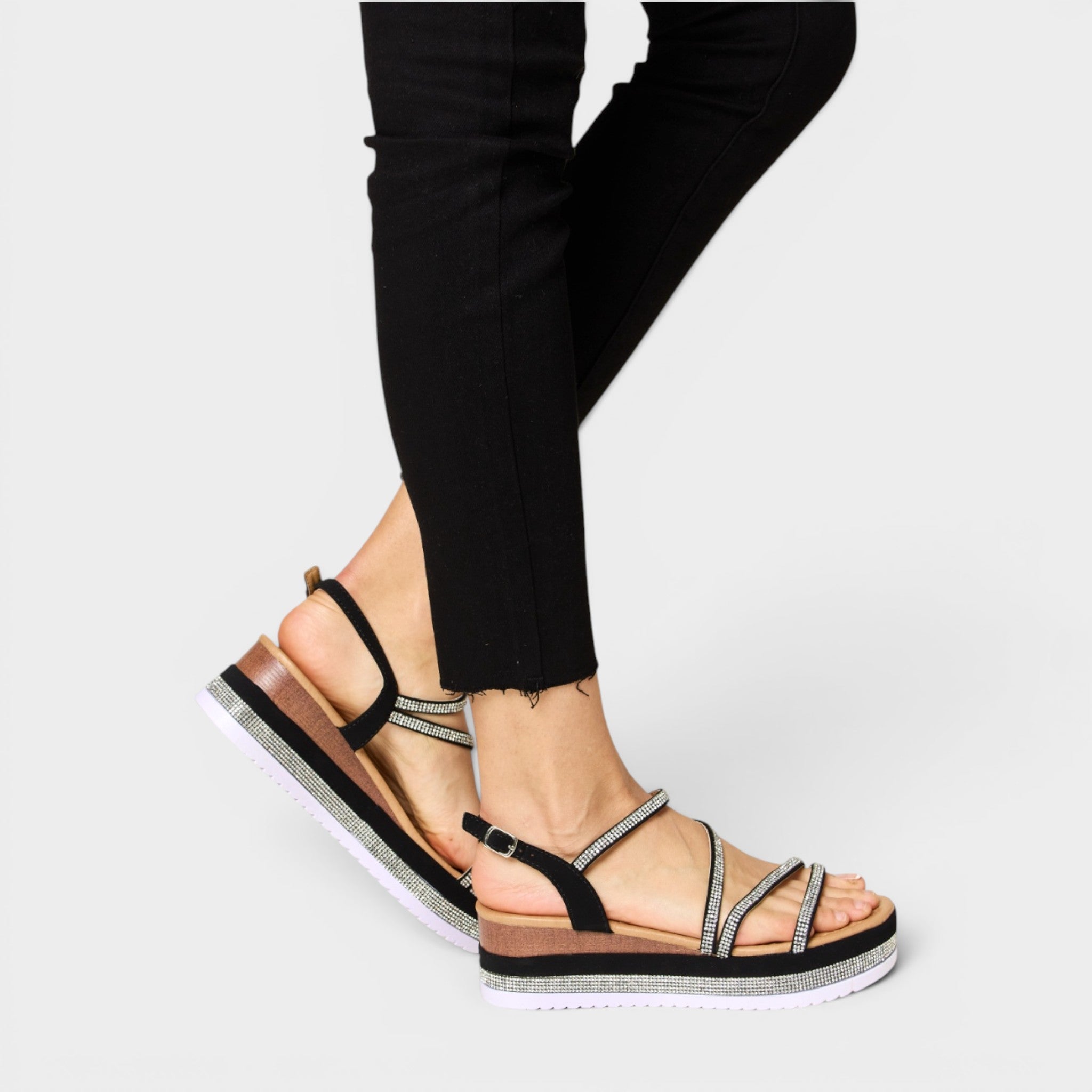 Black sandals with metallic straps worn with black pants on a light gray background