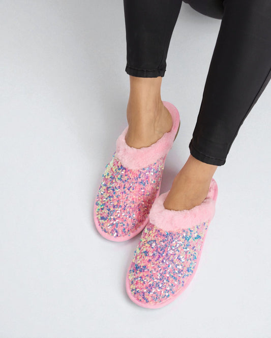 Pink slippers with multicolored glitter sole worn by a person on a light gray background