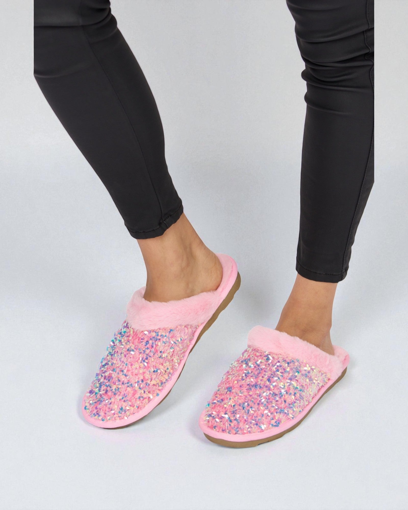 Pink slippers with colorful sequins worn by a person in black pants on a light gray background
